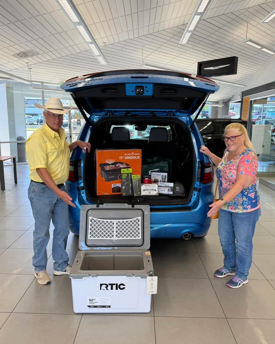 Two giveaway winners standing beside prizes in a dealership showroom.